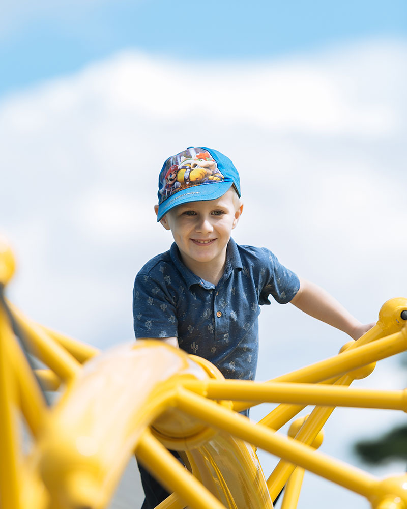 A young boy climbs to the top of a yellow steel playground structure, his head and shoulders are visible and he is smiling at the camera.